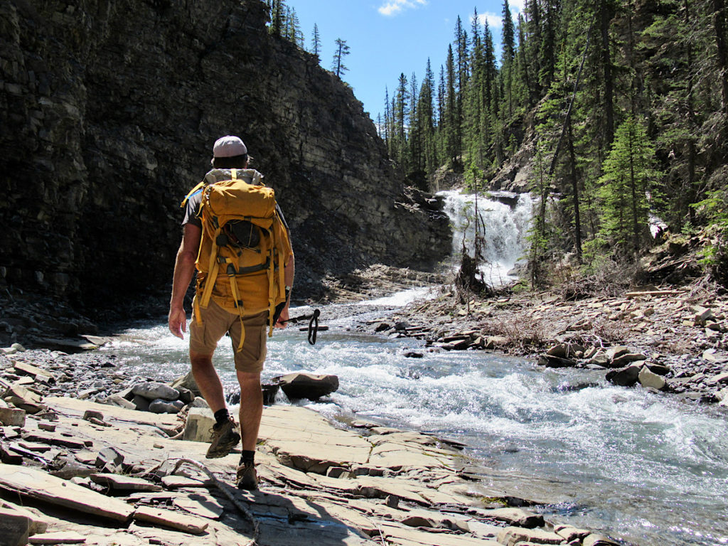 Man with big gold-coloured backpack walking on rock beside stream leading to small waterfall in a wooded environment.