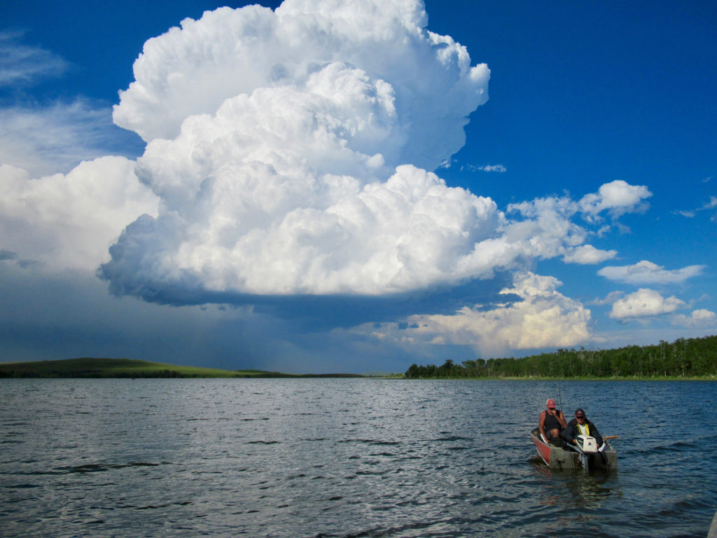 A small red aluminum boat with motor and two anglers heading out on lake after towering cloud mass has passed .