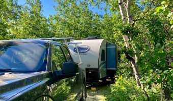 Gray truck and blue and white Rpod trailer with slide out park in treed campsite.