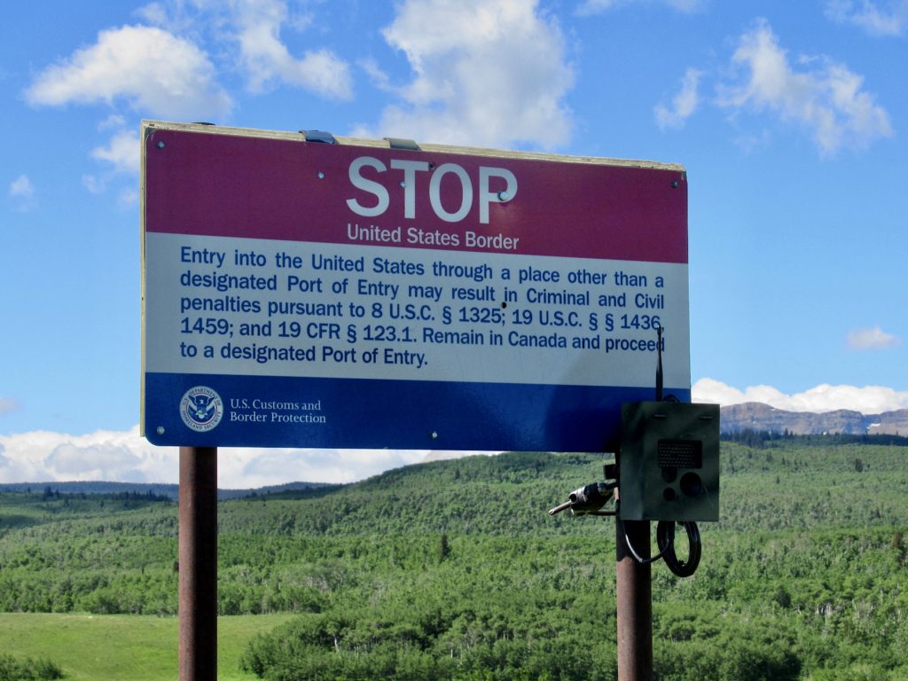 US border sign in red, white and blue with stop written in bold letters and explanation of crossing at proper border entry.