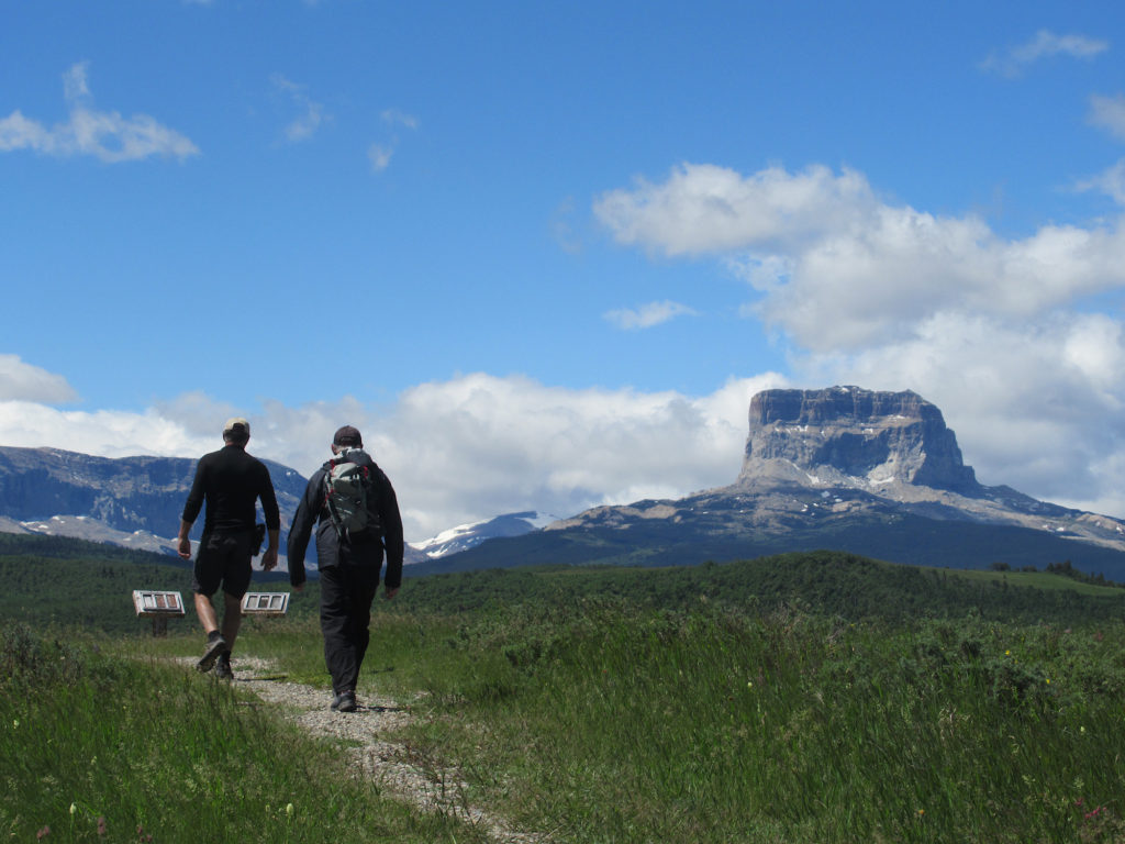Two men in dark clothes hiking on gravel path through grassy meadows with blocky grey mountain looming in distance under blue sky.