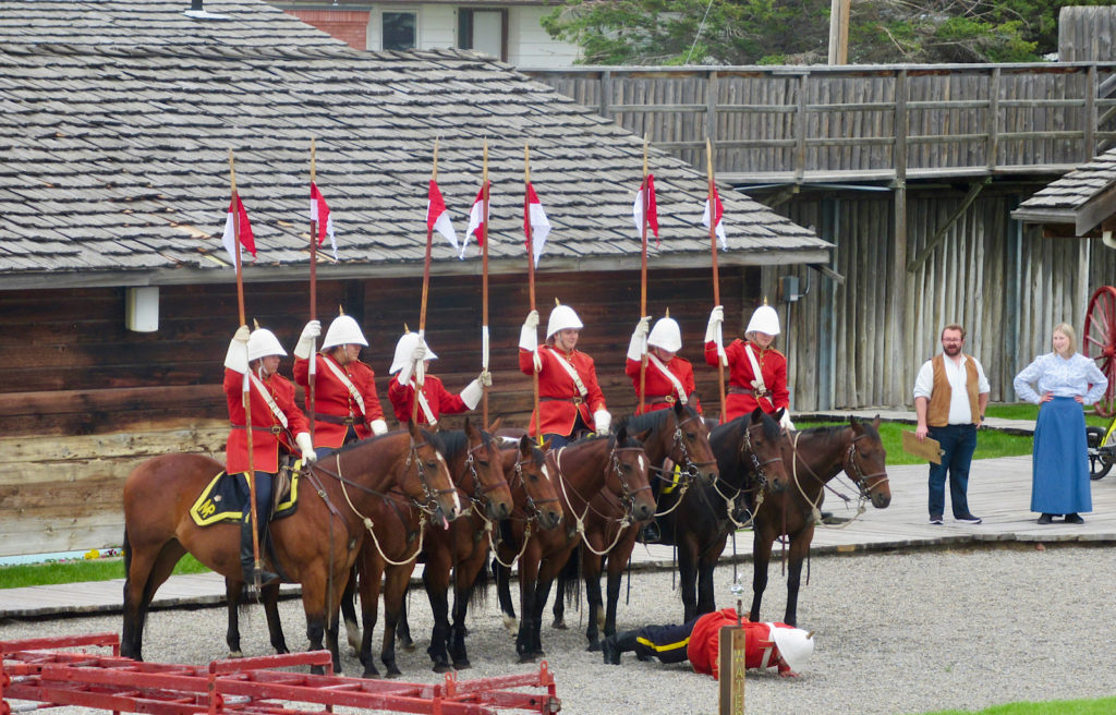 Six mounted horseback riders wearing red jackets and white hats and holding tall pole with red and white flags stand behind another dismounted rider doing pushups on gravel surface.