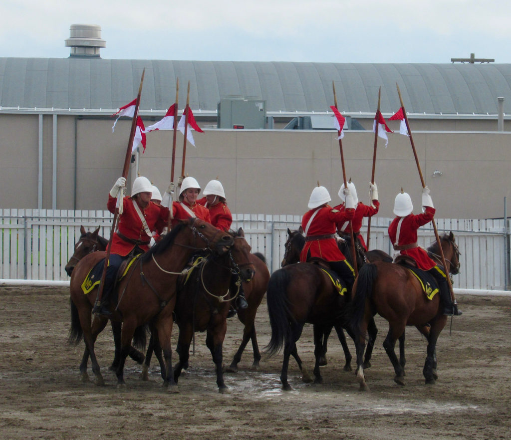 Pairs of mounted riders wearing red jacket and carrying a small flagpole circling like a carousel.