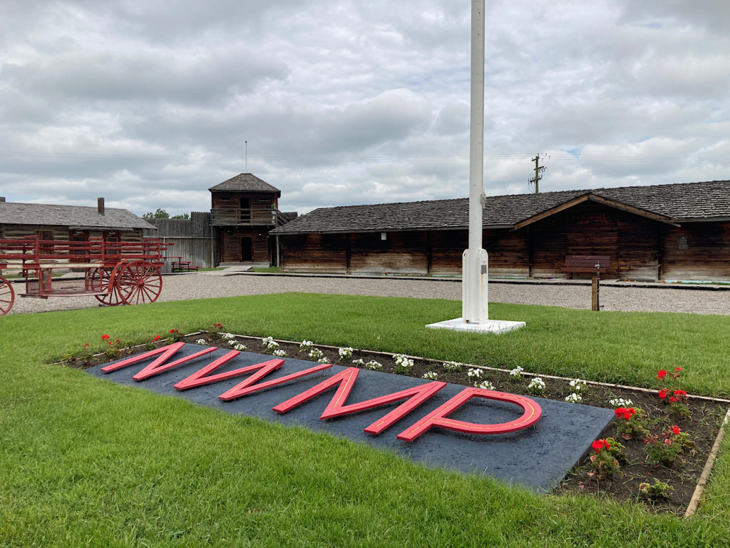 Large red letters spelling N-W-M-P in grassy area surrounded by wooden buildings.