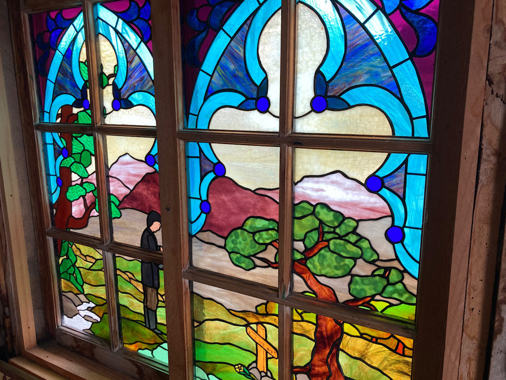 Stained glass window depicting a prairie scene with a person standing beside a grave underneath a tree.
