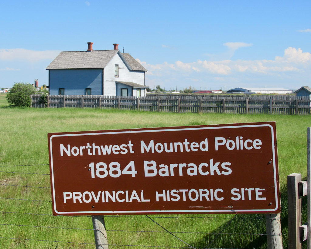 Brow sign with white lettering reading: "Northwest Mounted Police 1884 Barracks Provincial Historic Site" stands in front of grassy field with light blue building in background.
