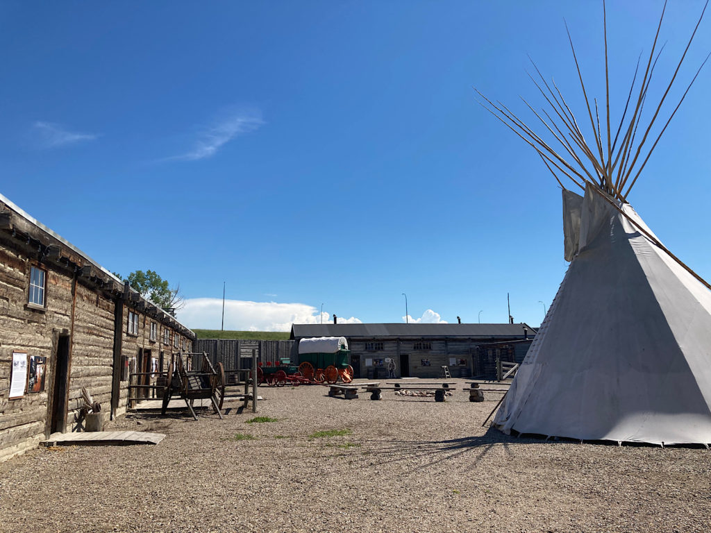Large open gravel area with a white teepee set up to one side and wooden buildings to left and in background.