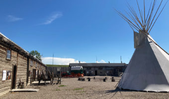 Large open gravel area with a white teepee set up to one side and wooden buildings to left and in background.