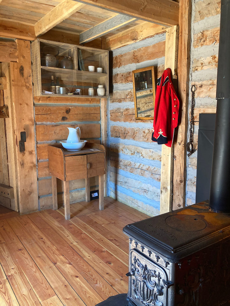 Interior of log cabin with cast iron stove, a red jacket hanging on the wall and a small table with ceramic water pitcher and bowl beneath open wood shelves.