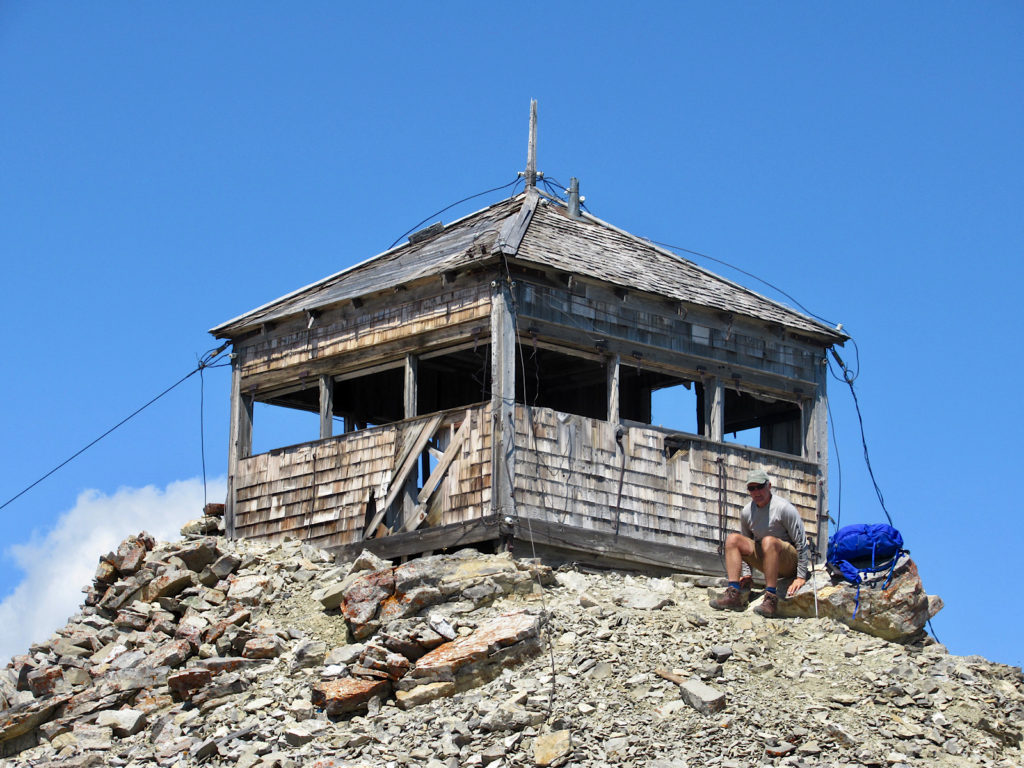 Weather worn small building on a rocky peak with man sitting on a rock with a blue backpack beside him.
