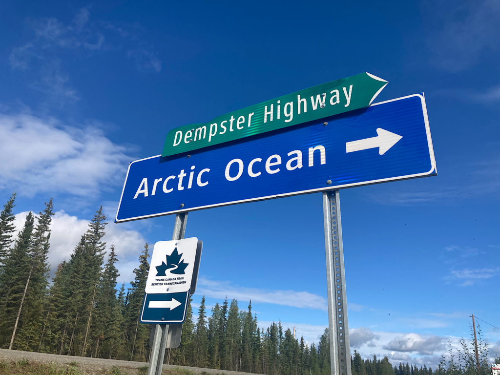 Road sign with Dempster Highway written in white letters on green background above Arctic Ocean written in white letters with white arrow pointing right on blue background.