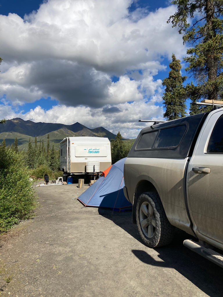 Muddy white truck with black canopy in campsite in front of blue tent and white trailer.
