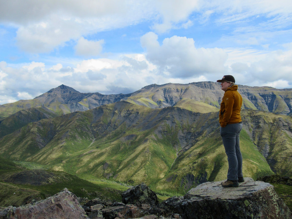 Woman in goldish-brown long sleeve shirt and grey pants standing on a rock to right side of picture with mountain ranges in back.