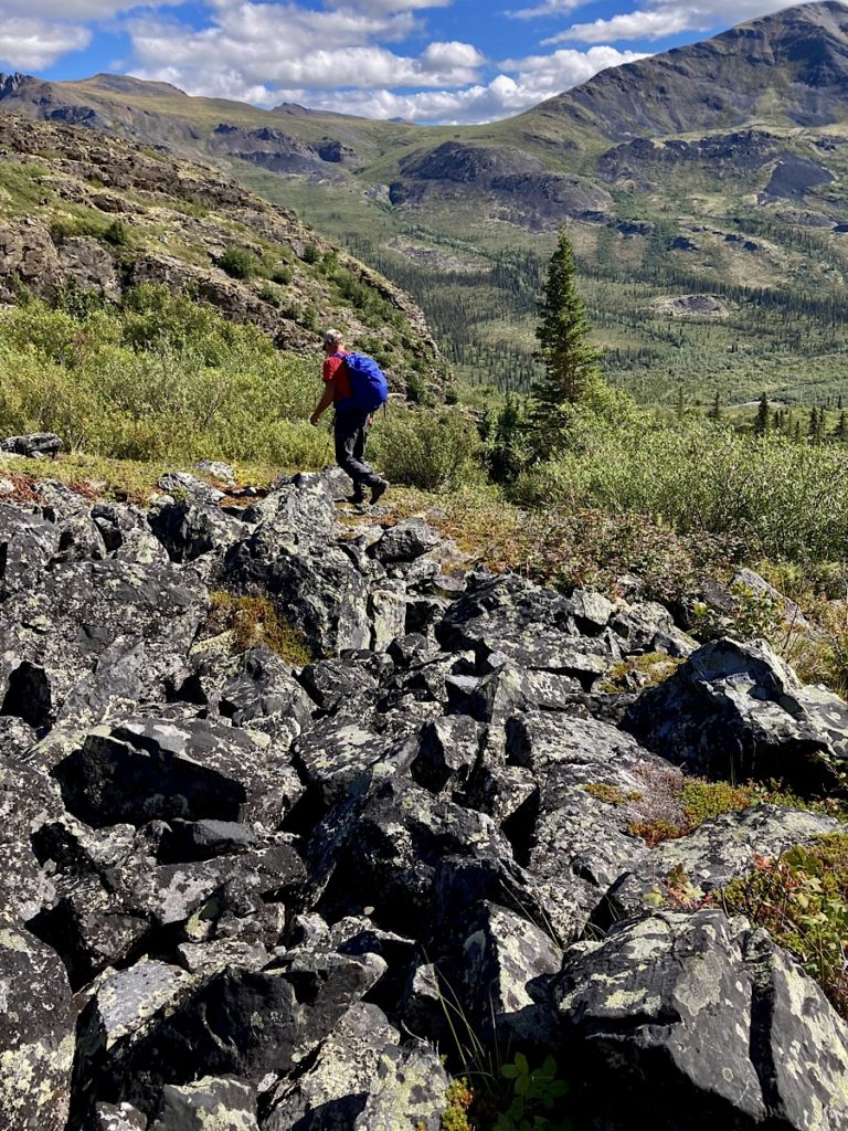 Man in red shirt and dark grey pants wearing blue backpack and hiking across a large boulder field.