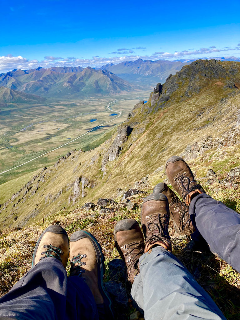 Three pairs of outstretched legs with leather hiking boots on feet in front of scenic mountain vista.