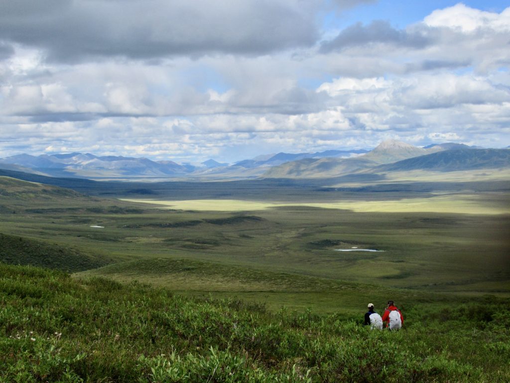 Two hikers in distance walking through tundra in wide open green valley.