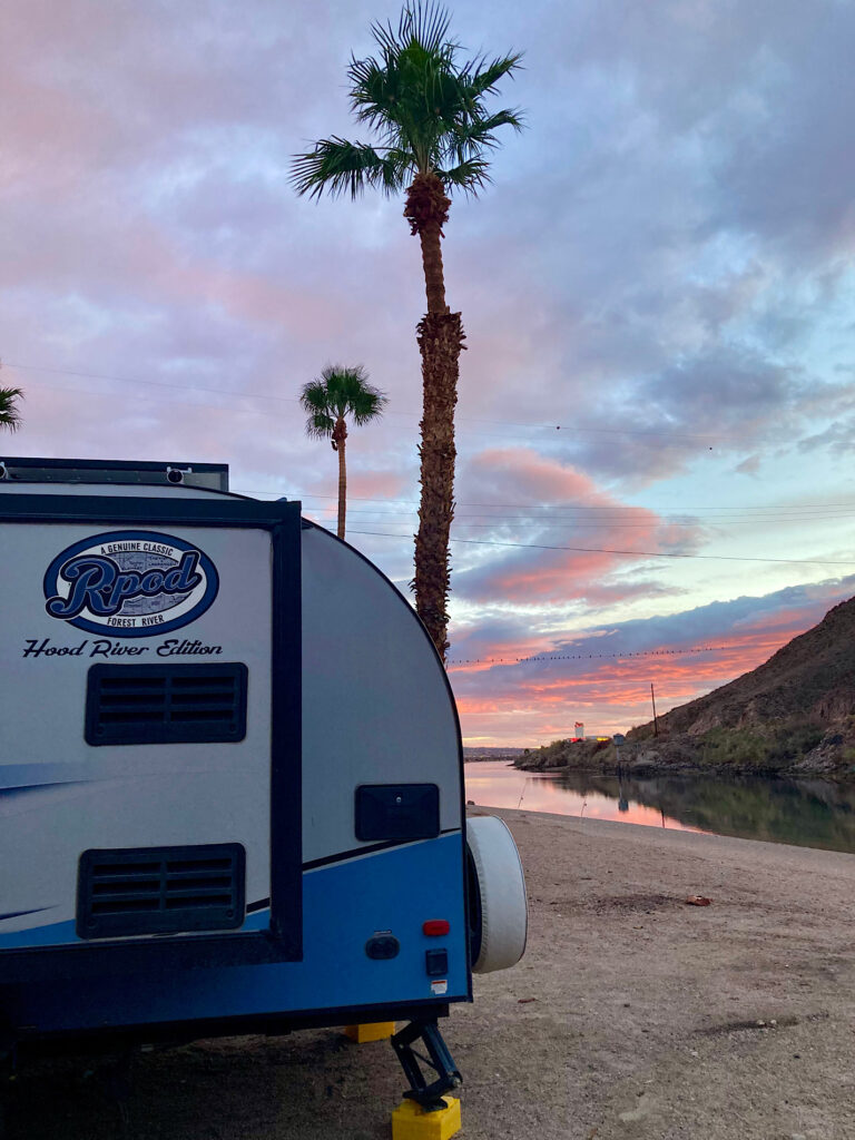Rpod Hood River Edition blue and white trailer set up near palm tree beside river with pink sunset above.