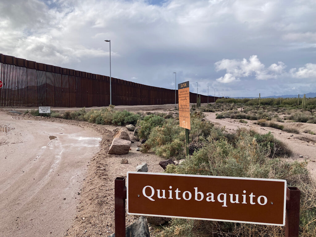 Brown sign with white lettering reading: Quitobaquito. In background is long, tall metal fence.
