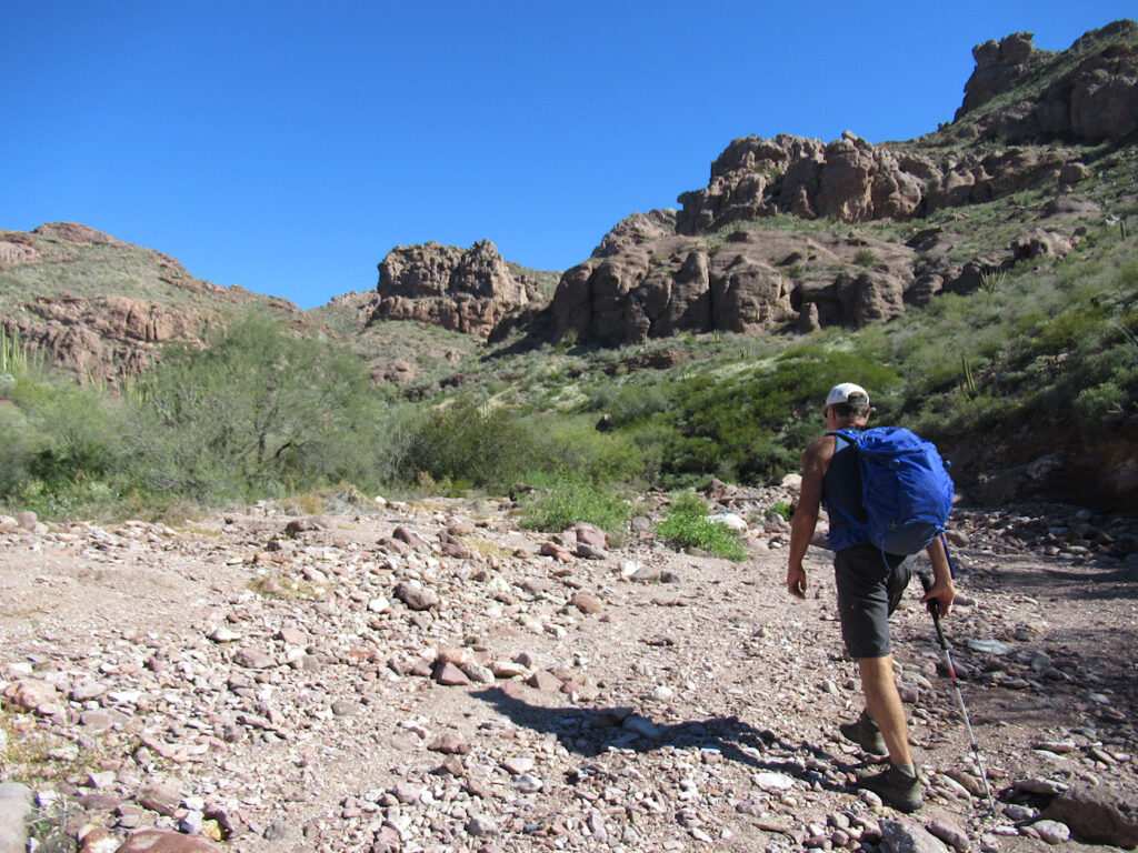 Man in shorts carrying blue backpack heading up rocky wash with distant cliffs under blue sky.