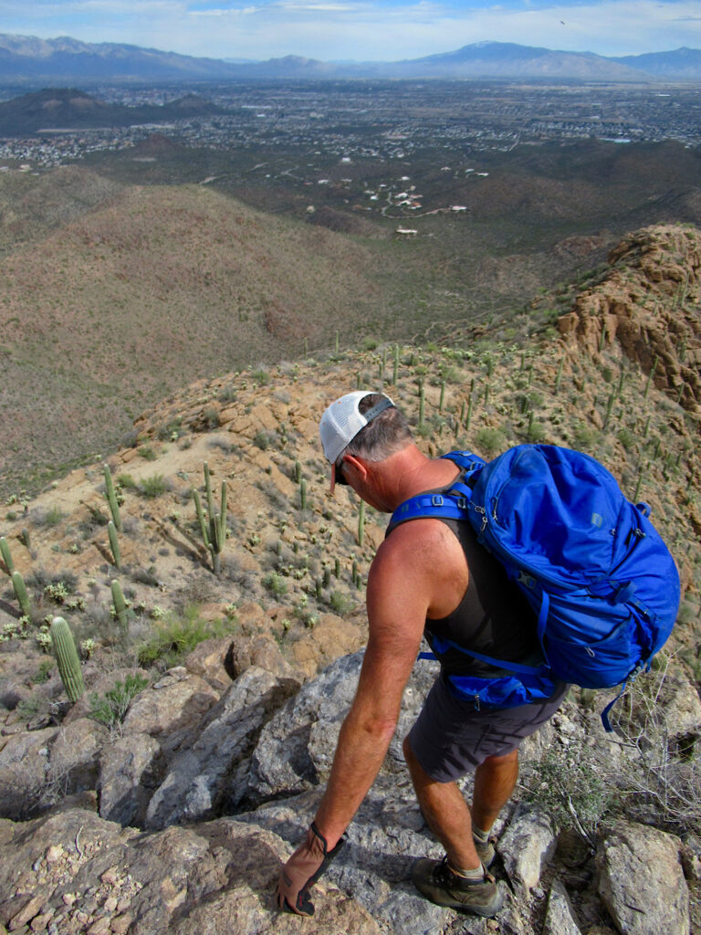 Man in short sleeve top and shorts wearing ball cap and carrying blue pack climbing down a steep mountain.