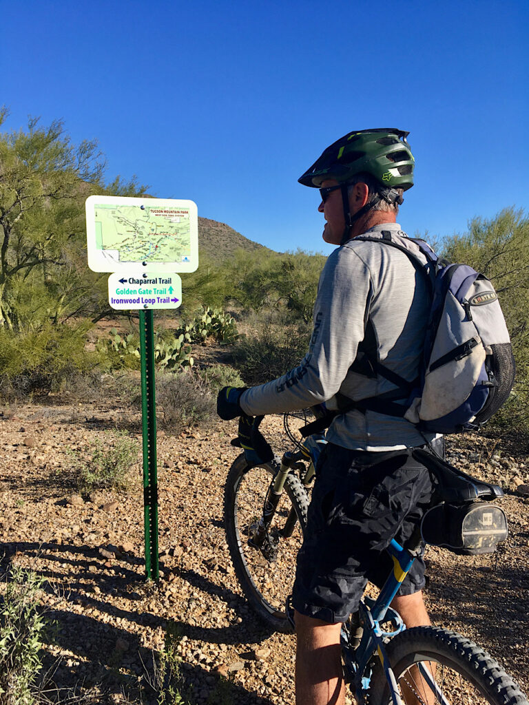 Man in long sleeve grey shirt and shorts wearing bicycle helmet and small backpack standing over his bike and looking at a trail sign.