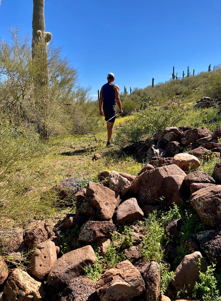 Man in shorts and short sleeve shirt walking away from brown pile of boulders under blue sky.