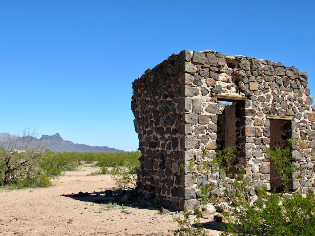 Old stone building with two window openings in flat gravel area with dark mountain in distance under blue sky.