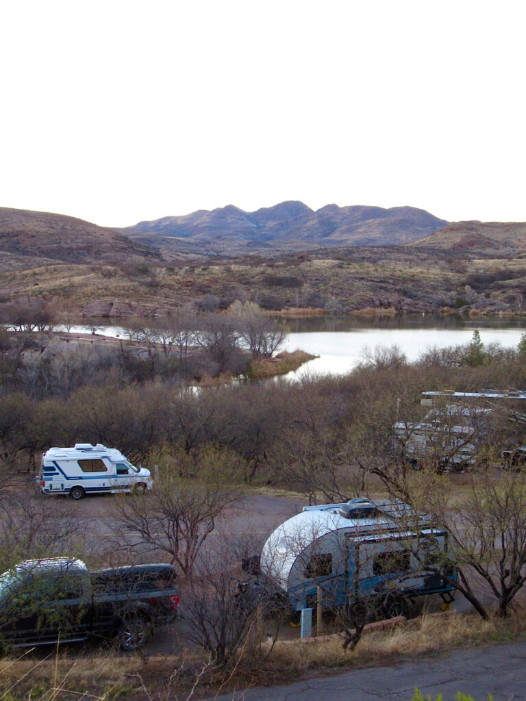Campground with a truck and trailer and distant RV in front of lake. 