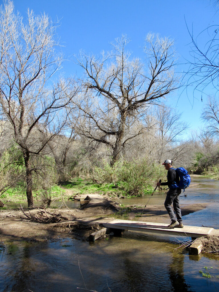 Man wearing blue backpack, long pants and coat walking across wooden bridge over small creek.
