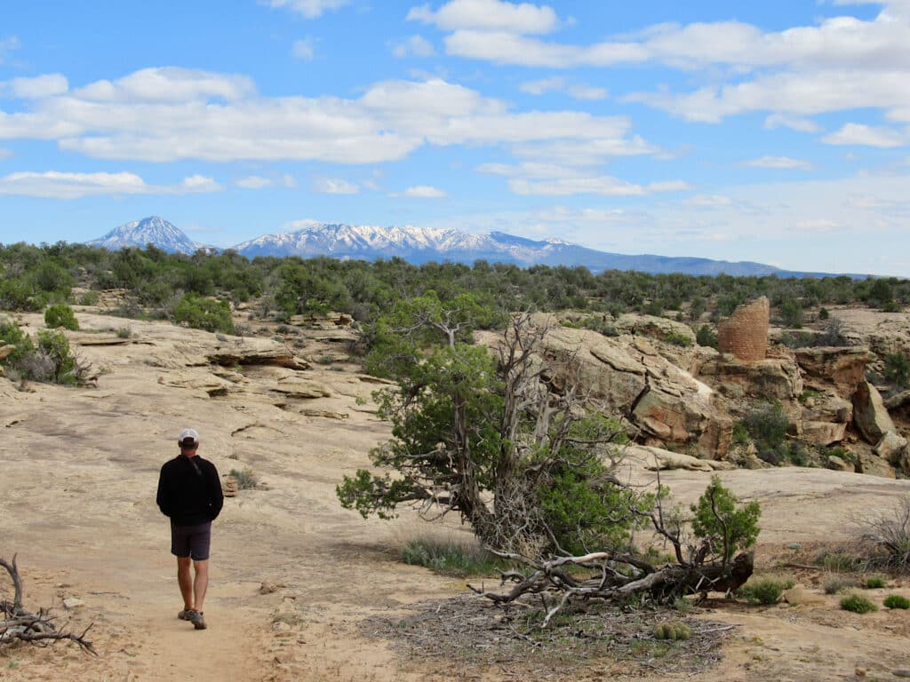 Man in long sleeve black shirt and shorts walking across open, light brown rock with reddish-brown, circular tower on edge of canyon in distance.