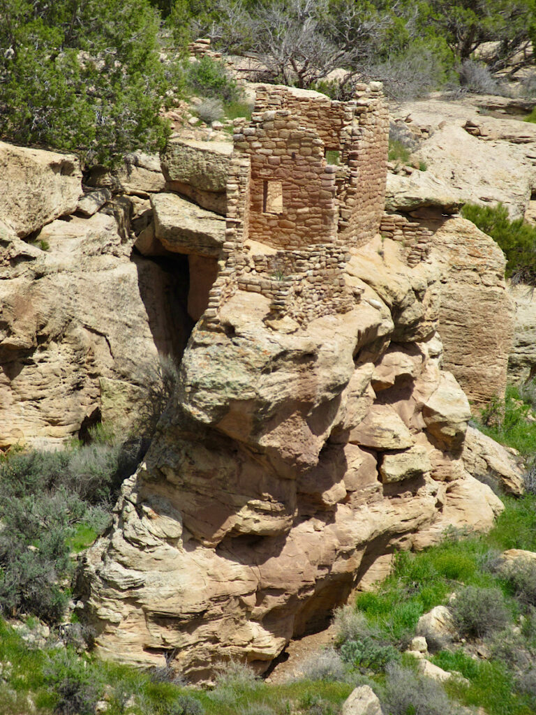 Square tower built on rugged, reddish-brown sandstone cliff edge high above green canyon floor.