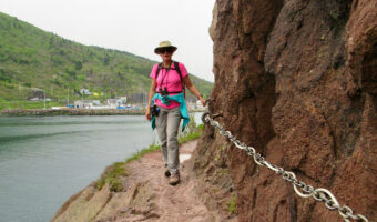 Woman in beige pants, pink top and widebrimmed hat holding onto a chain bolted to a rock wall on narrow rocky path above water.