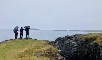 Three people in raingear, two with umbrellas, standing on green grassy cliff edge overlooking ocean.