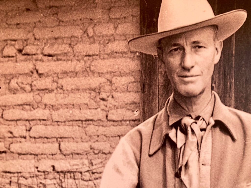 Sepia-toned image of a man wearing a cowboy hat, cowboy scarf, vest and long shirt in front of adobe brick building