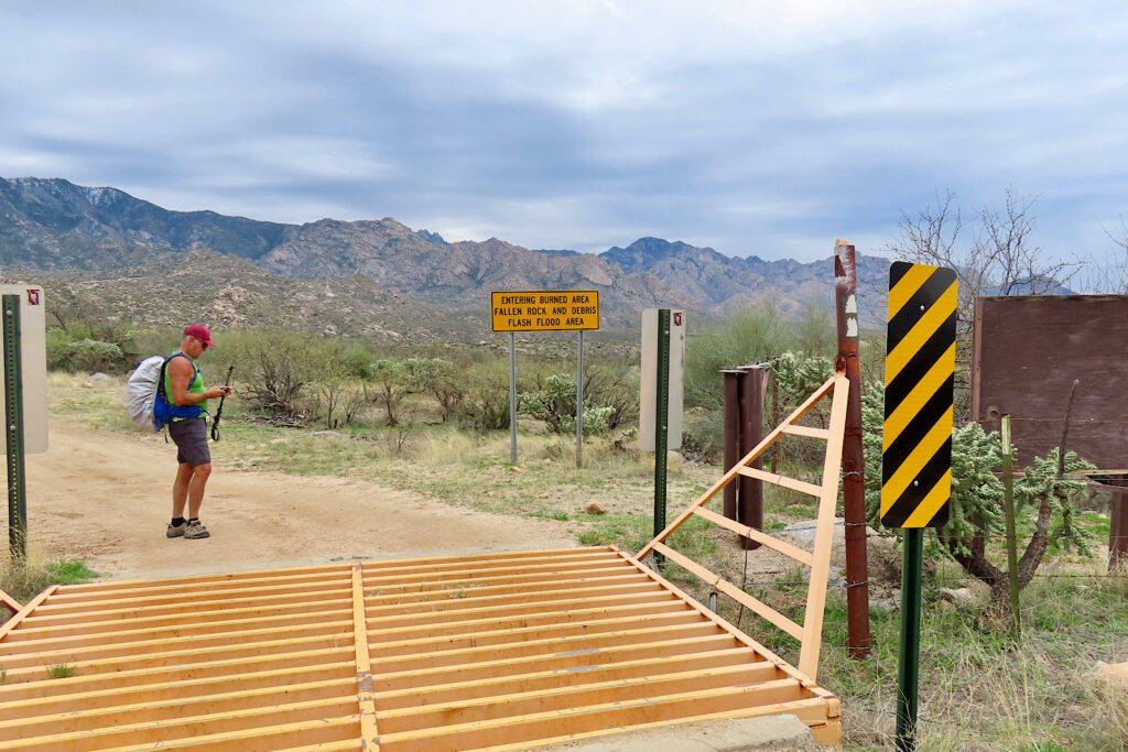 Man in shorts and short sleeved top wearing backpack standing beyond a yellow metal cattle guard with mountains in distance.