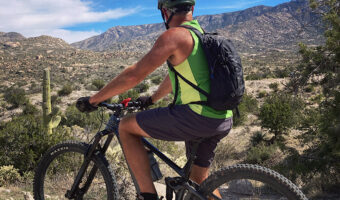Man in green short sleeved shirt and shorts with backpack on a bike overlooking light brown granite rocks.