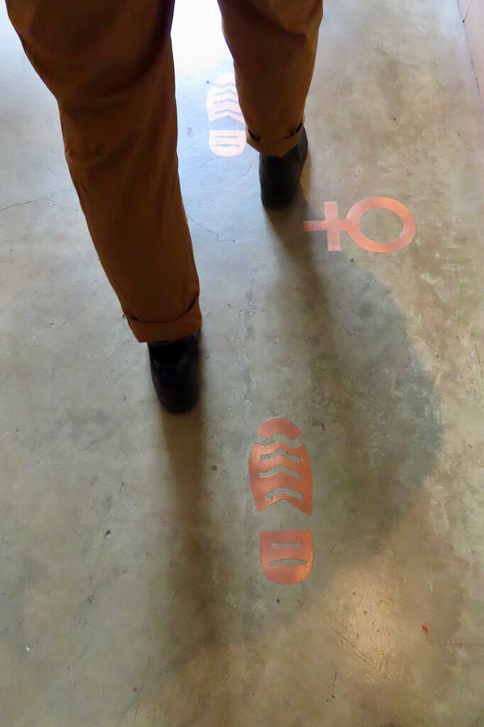 Woman in black boots walking on concrete floor beside inlaid copper footprints.