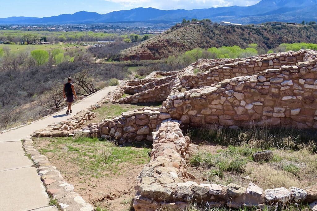 Man walking on sloping paved path beside rock ruins with distant mountains.