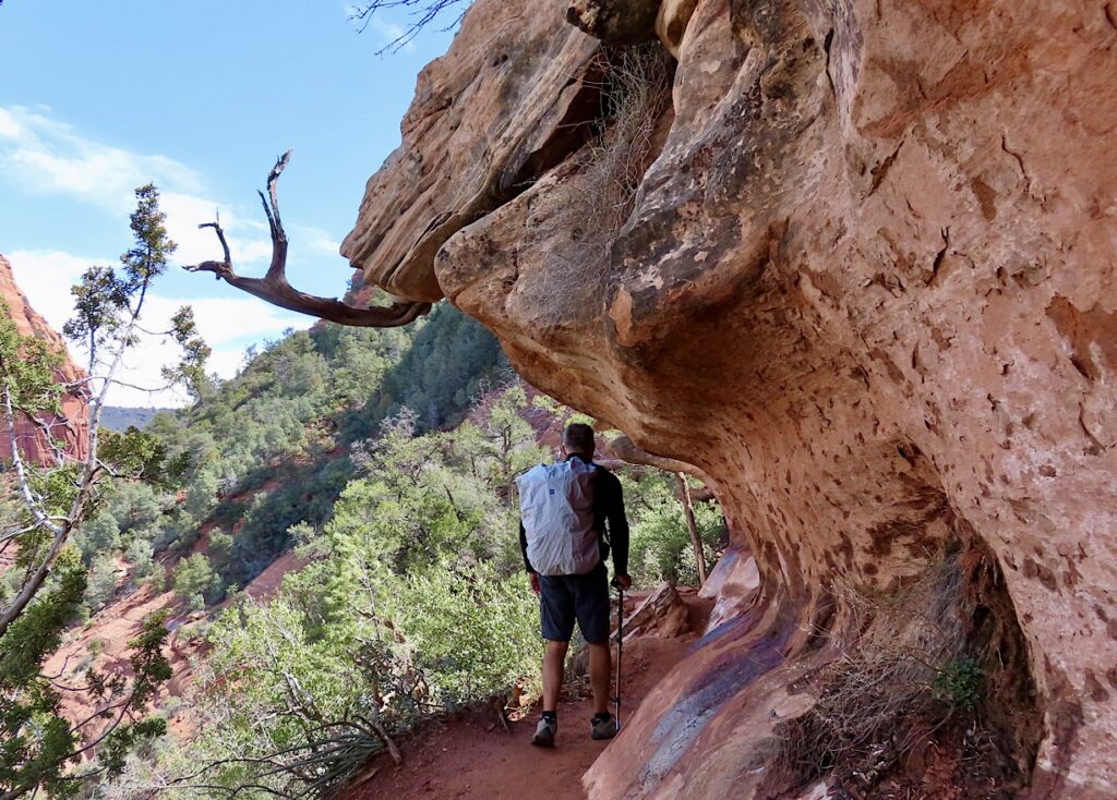 Man in shorts with grey pack walking on narrow path with overhanging reddish-brown sandstone cliffs.