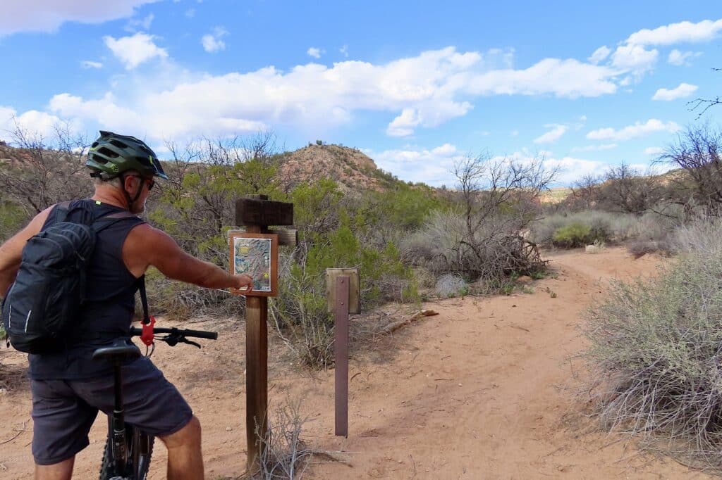 Man in black shirt and grey shorts wearing bike helmet and black pack on a bike pointing at trail sign.