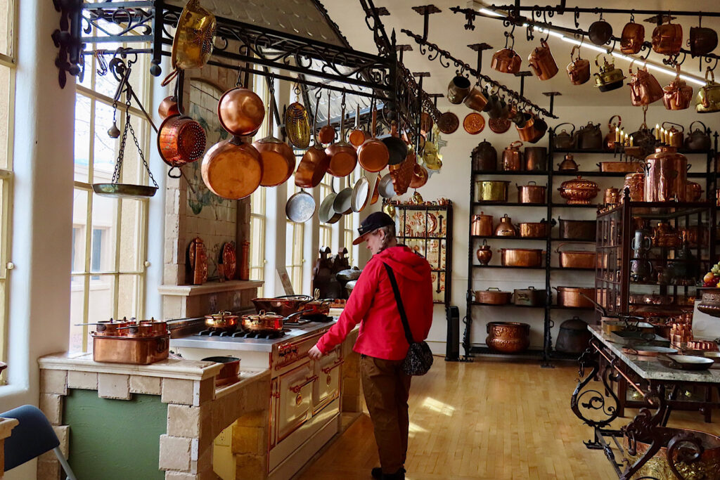 Woman in red coat, black hat and brown pants standing by old stove with copper pots handing all around and on shelves.