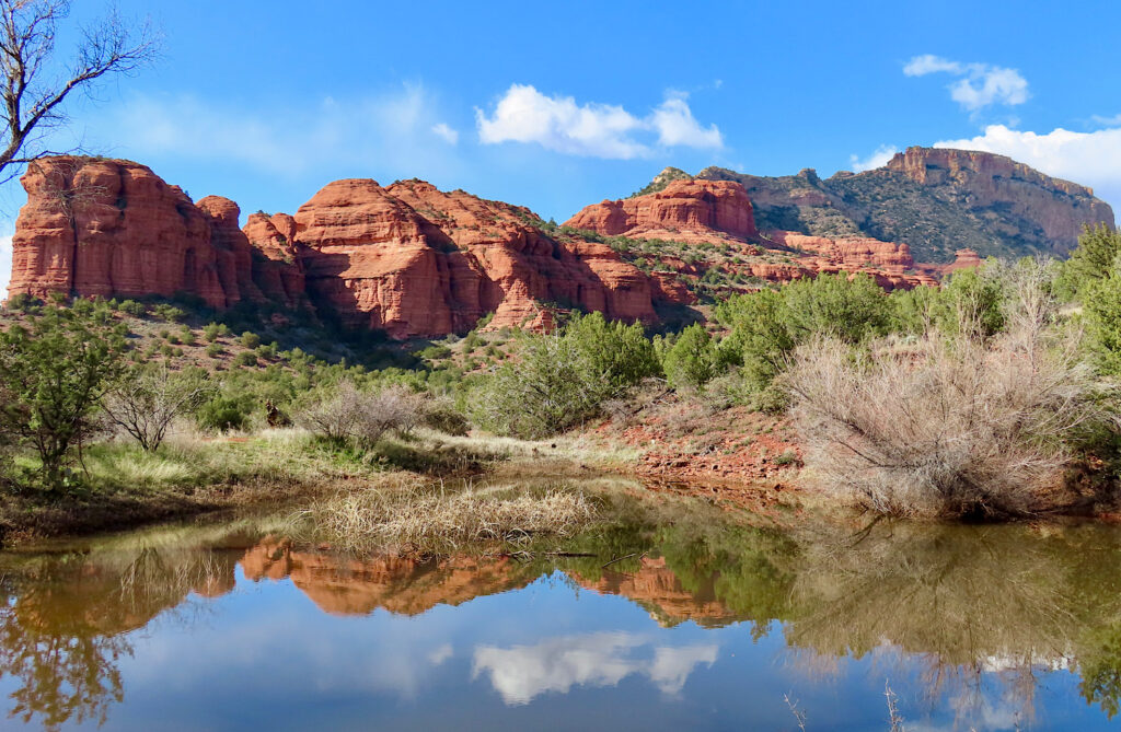 Red sandstone cliffs, blue sky, and green trees mirrored in small lake.