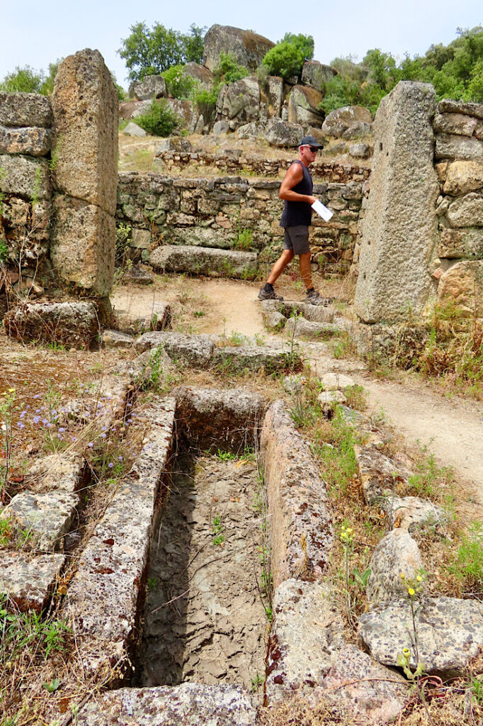 Man holding a white booklet walking in background of stone building ruins.