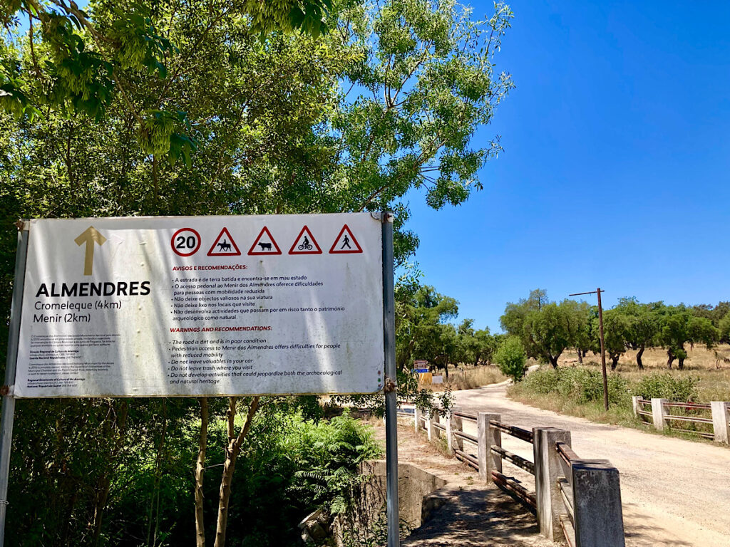 Route sign with warnings beside gravel road.