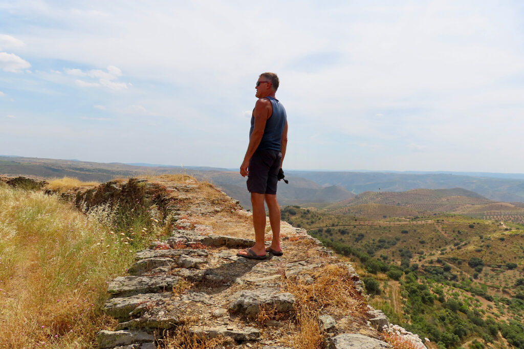 Man in dark shorts and short-sleeved shirt  walking old stone wall above green valley.