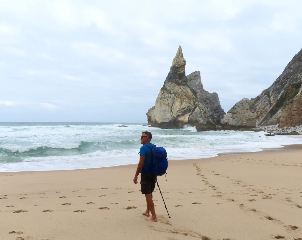 Man walking on brown sand beach with rolling waves and jagged rock outcrop behind.