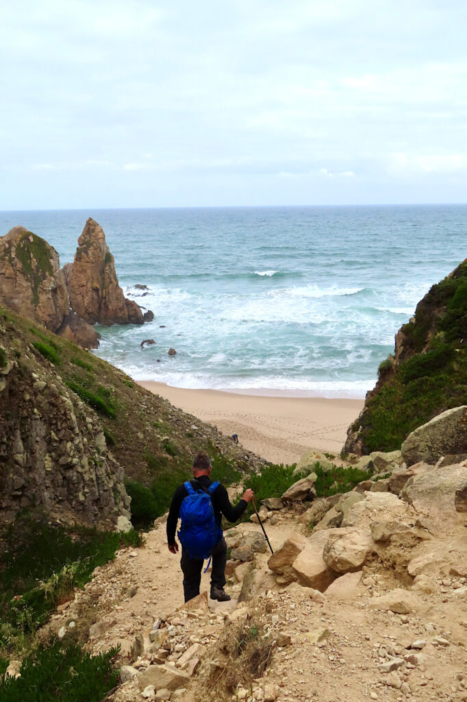 Man in black coat and pants with blue backpack heading down rocky trail to ocean and beach.