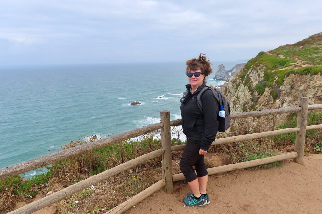 Woman in black coat and black pants wearing backpack and standing in front of fence with background cliffs down to the blue ocean.