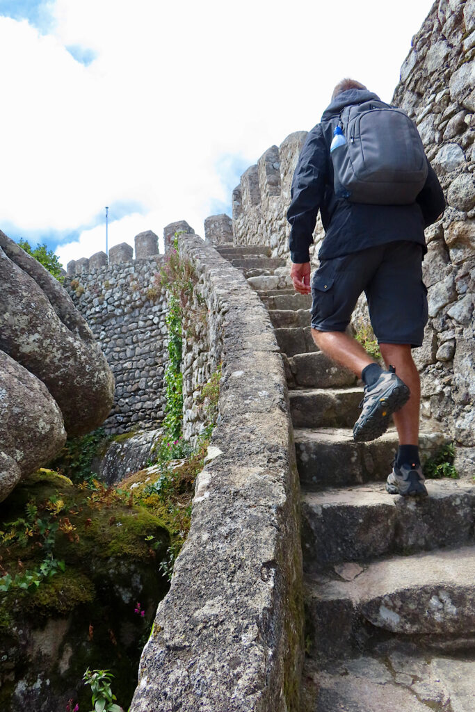 Man in dark coat and shorts wearing backpack climbing stone stairs up to a castle wall.