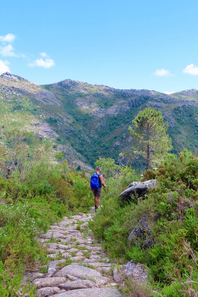 Man in shorts carrying a backpack and walking a stone path in a mountain valley under blue sky.